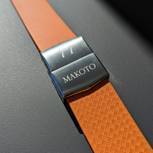Close-up of an orange watch strap with a metal clasp engraved 'MAKOTO'.