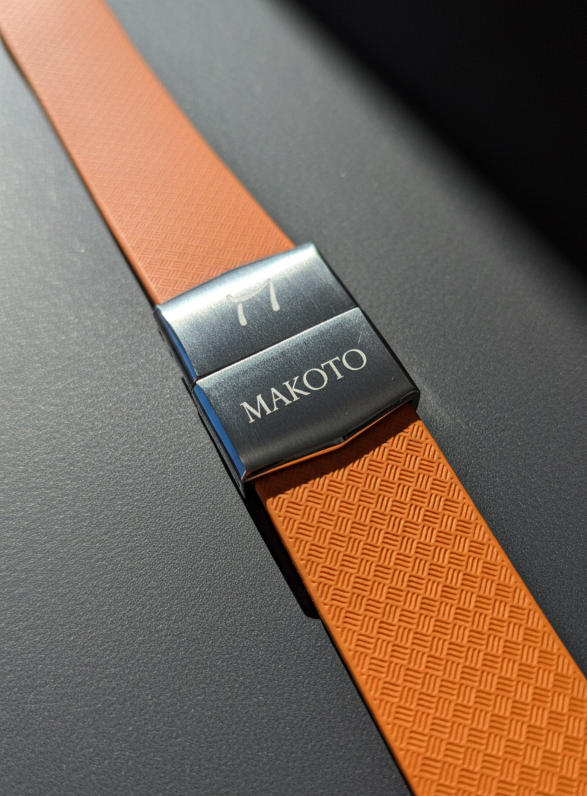 Close-up of an orange watch strap with a metal clasp engraved 'MAKOTO'.